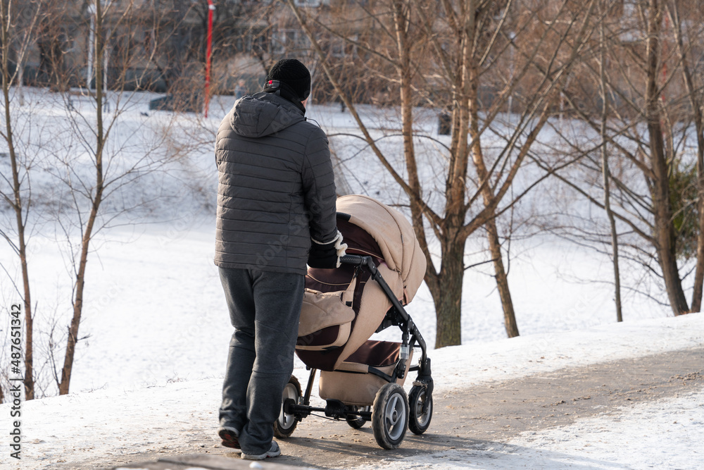 A young father walks with a baby in a stroller on a winter sunny day in a city park