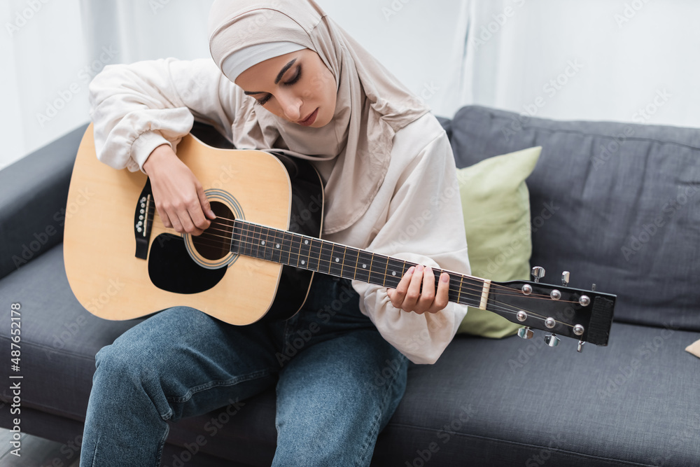 arabian woman in hijab playing guitar on couch at home.