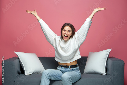 Overjoyed lucky young girl celebrating win with raised arms, screaming with euphoria, sitting on sofa