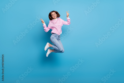 Photo of pretty excited woman wear striped sweater jumping up v-sings empty space isolated blue color background