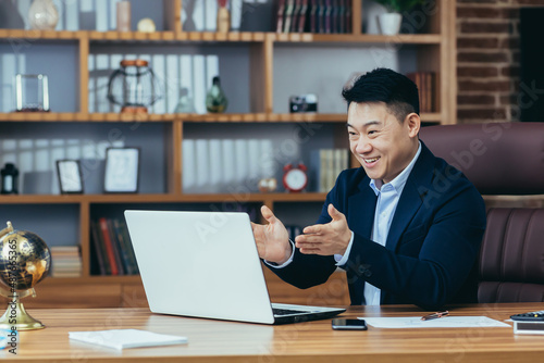 successful and happy asian boss looks at laptop screen and smiles, video call with colleagues, tells financial plans, online business meeting