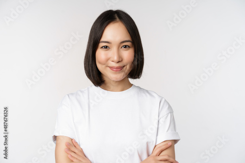 Close up portrait of confident korean girl, student looking at camera with pleased smile, arms crossed on chest, standing over white background