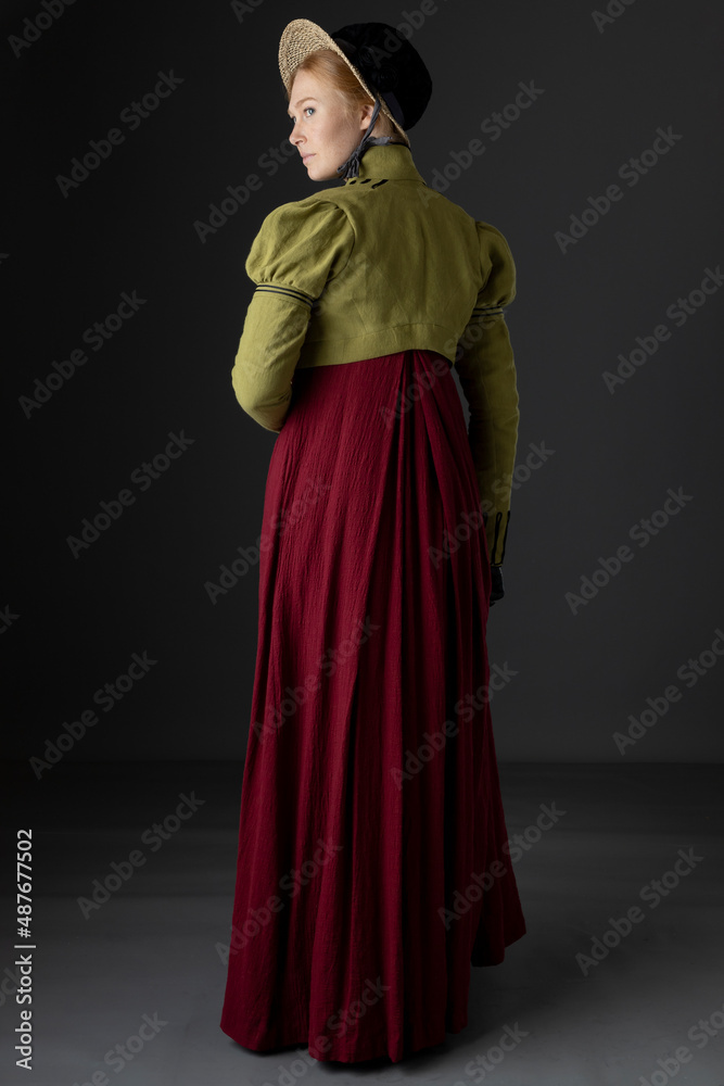 A Regency woman wearing a red day dress with a green linen spencer with black braid plus a bonnet and gloves against a studio backdrop A Regency woman wearing a red day dress with a green linen spencer with black braid plus a bonnet and gloves against a studio backdrop