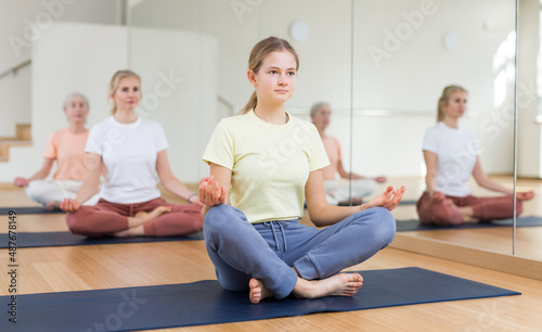 Teenage girl sitting on mat in lotus pose while training yoga in studio.
