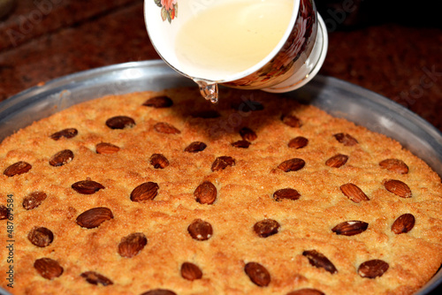 Pouring a mug of hot honey sugary syrup on a tray of basbosa with Almond , Basbusa is a middle eastern dessert that is usually baked in Ramadan month, selective focus of  Arabian sweet cuisine photo