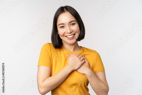 Romantic asian girfriend, holding hands on heart, smiling with care and tenderness, standing in yellow tshirt over white background photo