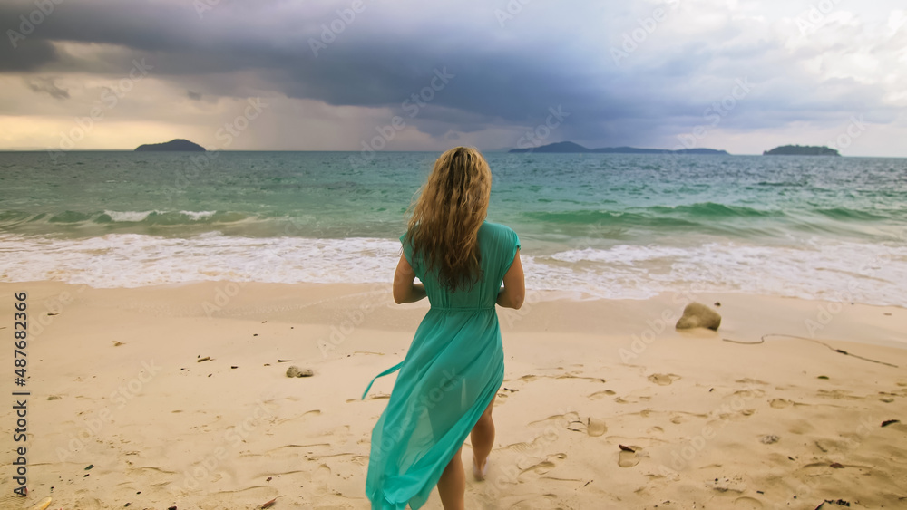 Beautiful long-haired woman walks to the stormy cloudy ocean on sand beach. Girl in blue swimsuit dress tunic. Concept rest in sea, tropical resort coastline traveling tourism summer beach holidays Beautiful long-haired woman walks to the stormy cloudy ocean on sand beach. Girl in blue swimsuit dress tunic. Concept rest in sea, tropical resort coastline traveling tourism summer beach holidays