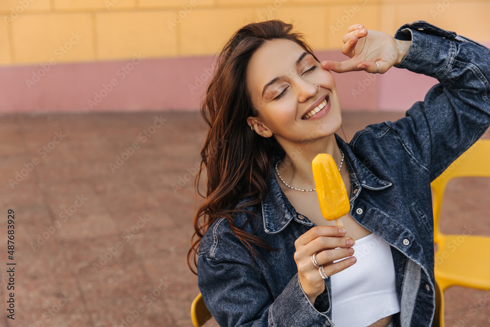 Modern caucasian girl sitting outdoors eating ice cream on warm summer day. Beauty with dark hair, eyes closed, wearing white top and denim jacket. Summer playful mood concept Modern caucasian girl sitting outdoors eating ice cream on warm summer day. Beauty with dark hair, eyes closed, wearing white top and denim jacket. Summer playful mood concept