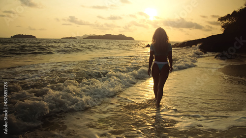 Silhouette woman walking alone on beach coastline, relax warm gold sunset. Woman walking on water in blue swimsuit and sunglasses. Concept rest tropical resort traveling tourism happy summer holidays