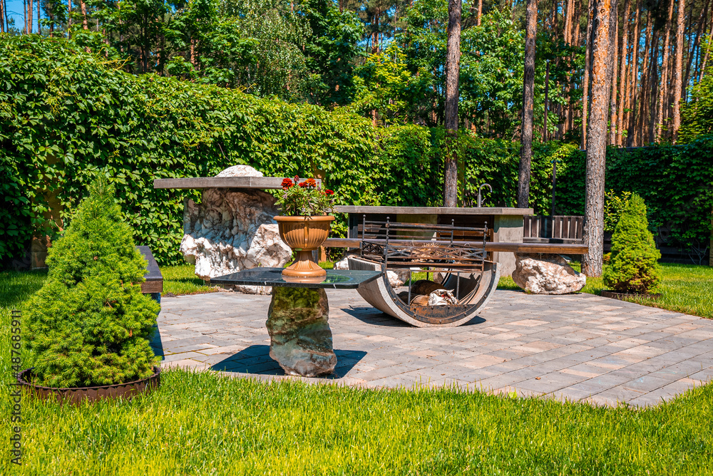 Obraz premium Beautiful sunny backyard with iron fire pit and bench on stone pavement, Garden rest area with fire pit surrounded by green shrubs