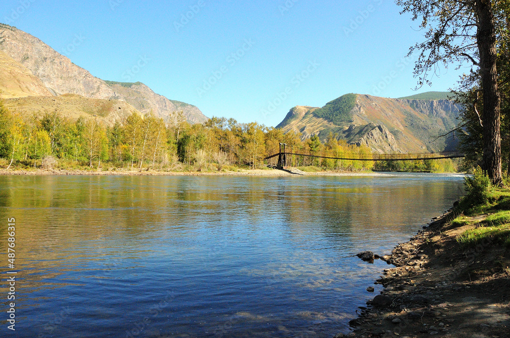 The calm expanse of a mountain river and a suspension bridge across it, surrounded by high mountains.