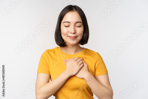 Romantic asian girfriend, holding hands on heart, smiling with care and tenderness, standing in yellow tshirt over white background photo