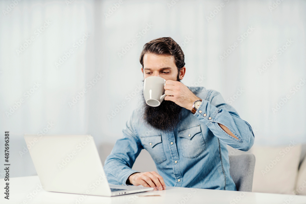 A bearded entrepreneur drinking his coffee and looking at laptop at home. A bearded entrepreneur drinking his coffee and looking at laptop at home.