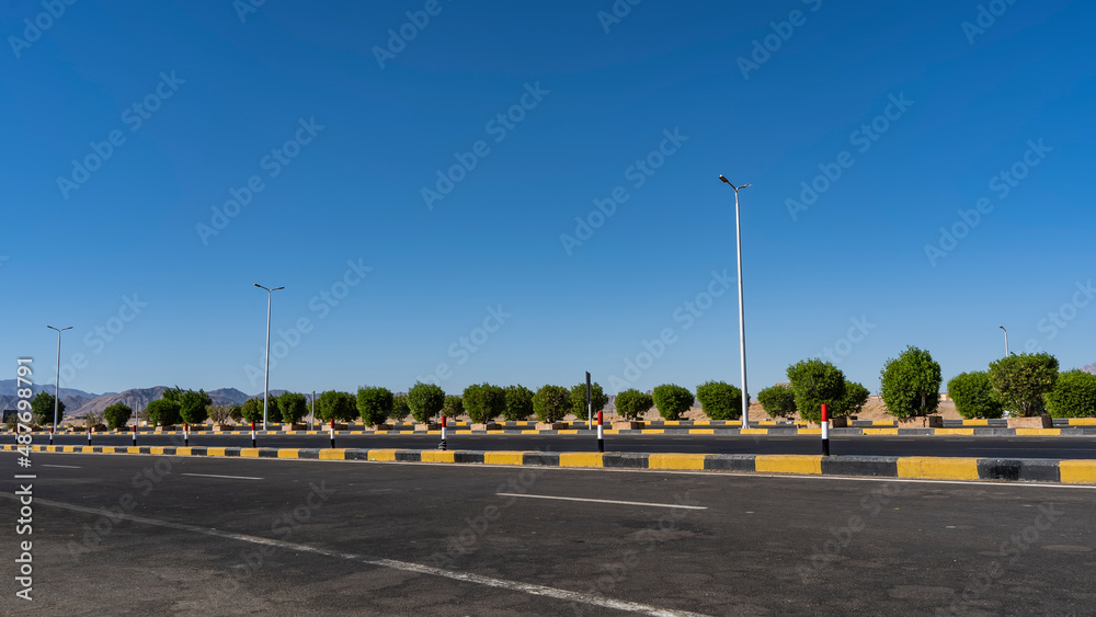 The highway is covered with new asphalt. The marking lines and the dividing barrier are visible. A row of green bushes along the road. Mountains against the blue sky in the distance. Egypt. Safaga