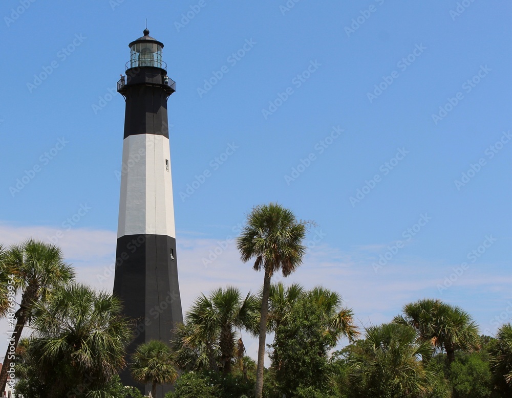 Scenic tall white and black lighthouse with searchlight amidst green palm trees, blue sky, and white clouds. Scenic tall white and black lighthouse with searchlight amidst green palm trees, blue sky, and white clouds.