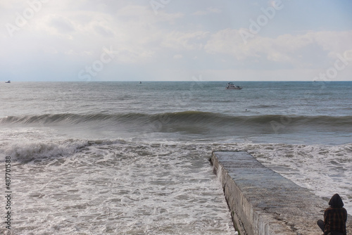 Storm waves roll off a concrete breakwater on which a hooded girl sits. Yachts at sea are fighting the storm. Seagulls fly over the waves. Selective focus.