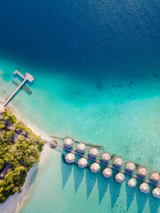 Overwater villas and white sand beach on tropical island for holidays vacation travel and honeymoon. Luxury resort hotel in Maldives or Caribbean with turquoise sea water. Drone aerial top down view.