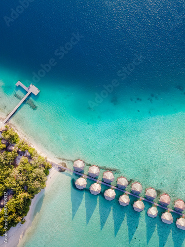 Overwater villas and white sand beach on tropical island for holidays vacation travel and honeymoon. Luxury resort hotel in Maldives or Caribbean with turquoise sea water. Drone aerial top down view.