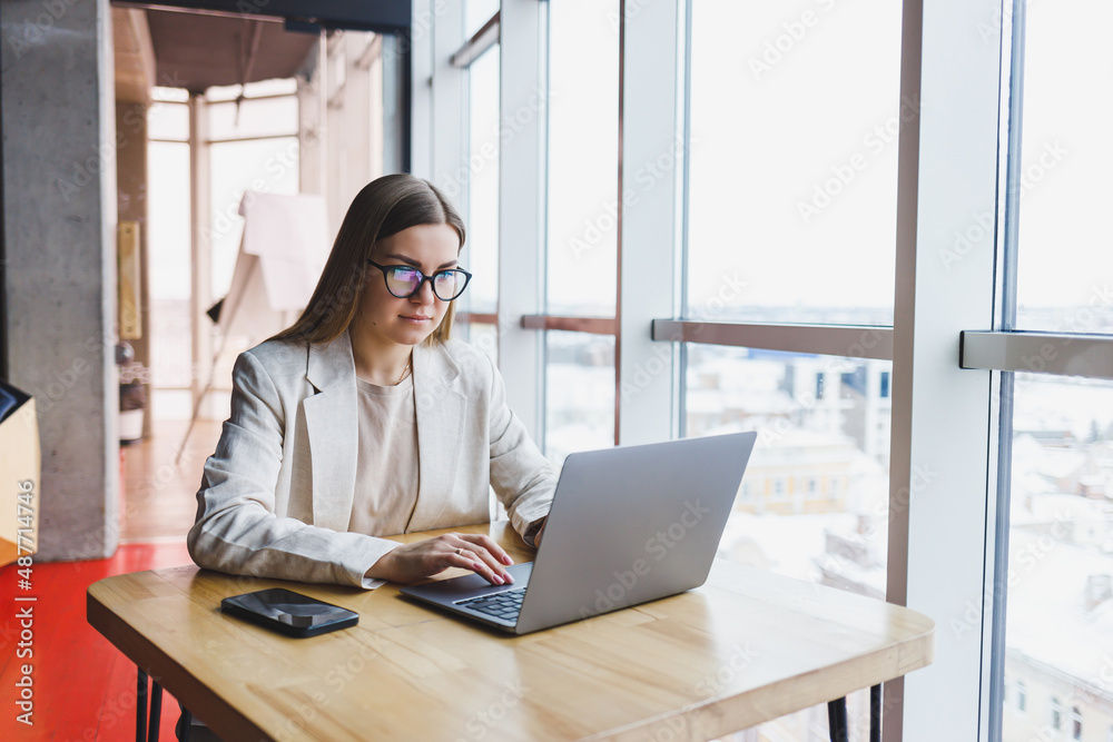 Image of a happy young woman in a jacket smiling and working on a laptop while talking on the phone in a modern office with large windows. Remote work Image of a happy young woman in a jacket smiling and working on a laptop while talking on the phone in a modern office with large windows. Remote work