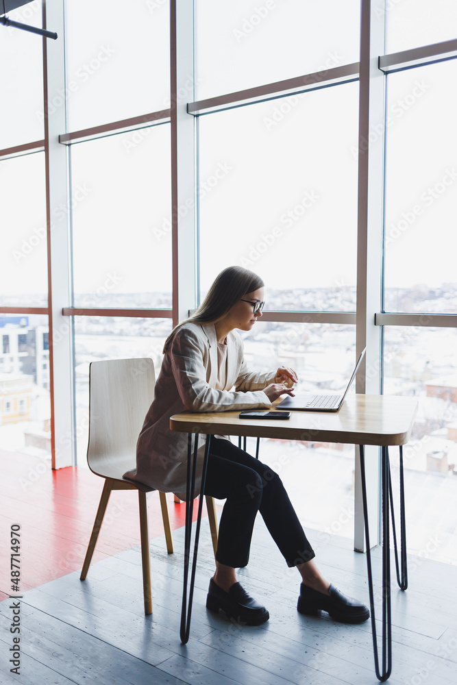 Image of a happy young woman in a jacket smiling and working on a laptop while talking on the phone in a modern office with large windows. Remote work Image of a happy young woman in a jacket smiling and working on a laptop while talking on the phone in a modern office with large windows. Remote work