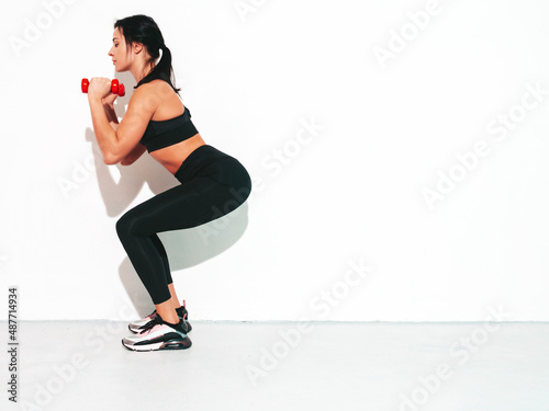 Fitness confident woman in black sports clothing. Sexy young beautiful model with perfect body. Female isolated on white wall in studio. Stretching out before training.Making squats with dumbbells
