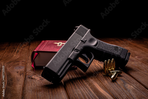 A pistol lies on a wooden table, resting on a bible in a red leather cover, soft focus