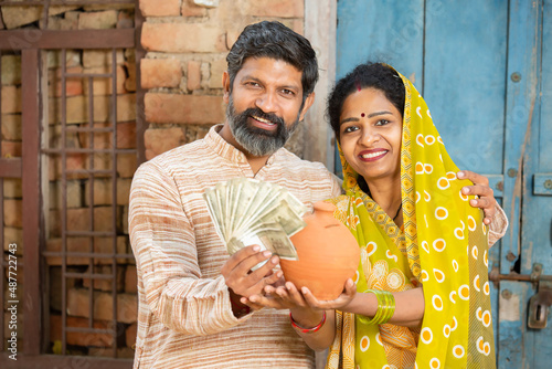 Portrait of happy rural couple holding indian rupee notes and clay money box or gullak. traditional piggy bank, save money, Investment and banking photo