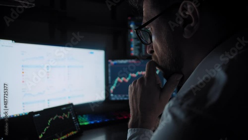 A bearded trader with glasses sitting near to computer monitors and analyzes the stock market date a close-up slow motion