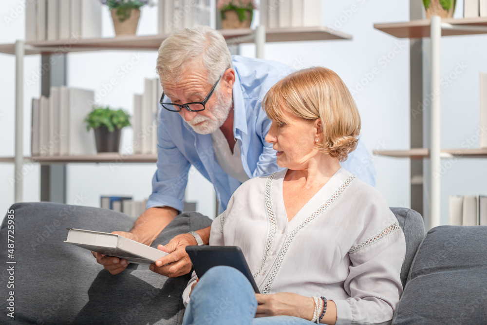 Fototapeta premium Portrait of happy senior couple in living room, mature woman and a man with a book and tablet on cozy sofa at home, Happy family concepts