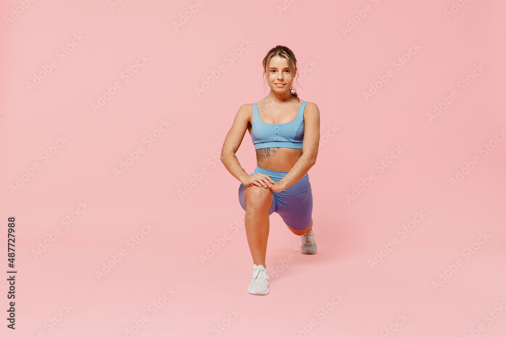 Full length young sporty athletic fitness trainer woman wear blue tracksuit spend time in home gym train do stretch legs squat exercise isolated on plain light pink background. Workout sport concept. Full length young sporty athletic fitness trainer woman wear blue tracksuit spend time in home gym train do stretch legs squat exercise isolated on plain light pink background. Workout sport concept.