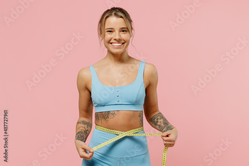 Young fun strong sporty athletic fitness trainer instructor woman wear blue tracksuit spend time in home gym hold measure tape on waist isolated on pastel plain pink background. Workout sport concept.