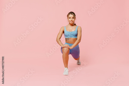 Full length young sporty athletic fitness trainer woman wear blue tracksuit spend time in home gym train do stretch legs squat exercise isolated on plain light pink background. Workout sport concept.