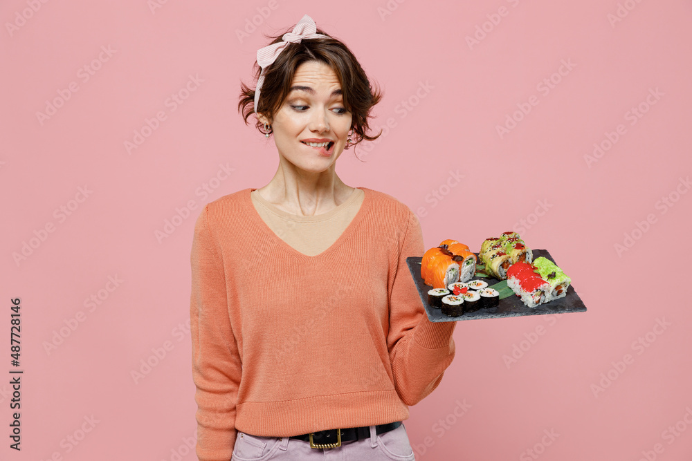 Young amazed impressed fun caucasian woman 20s in casual clothes hold in hand looking at makizushi sushi roll served on black plate traditional japanese food isolated on plain pastel pink background Young amazed impressed fun caucasian woman 20s in casual clothes hold in hand looking at makizushi sushi roll served on black plate traditional japanese food isolated on plain pastel pink background
