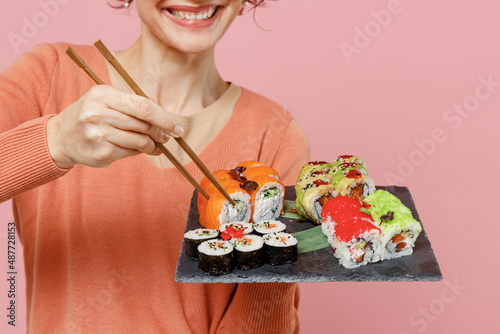 Cropped close up young smiling woman in casual clothes hold in hand makizushi sushi roll served on black plate traditional japanese food eat with chopsticks isolated on plain pastel pink background