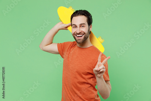 Young smilign happy cool man 20s in casual orange t-shirt hold yellow scateboard pennyboard show v-sign isolated on plain pastel light green color background studio portrait. People lifestyle concept. photo