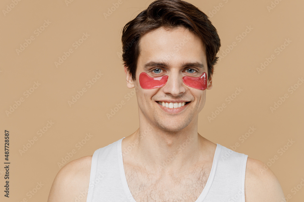 Attractive smiling young man 20s perfect skin in undershirt wearing gold patch under eye isolated on pastel pastel beige background studio portrait. Skin care healthcare cosmetic procedures concept.