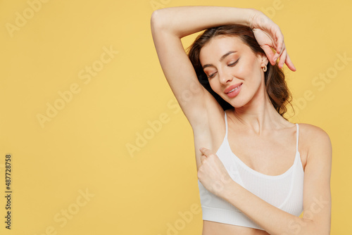 Satisfied tender european young brunette woman 20s in white brassiere underwear raise up hand show soft perfect shaved armpit isolated on plain yellow background studio Female beauty bodycare concept