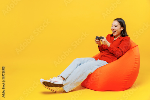 Full size body length fancy young woman of Asian ethnicity 20s wear casual clothes sit in bag chair hold in hand play pc game with joystick console isolated on plain yellow background studio portrait photo