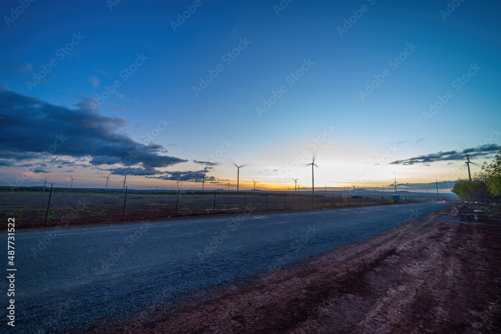 Fototapeta premium Wind turbine farm and agricultural fields on a summer day. Wind electricity generator in sunset sky, power plant, wind efficiency.