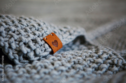 Warm knitted gray blanket, leather tag with image of heart, love. Soft, warm, handmade plaid. Texture for background or illustrations. Select focus, copy space.