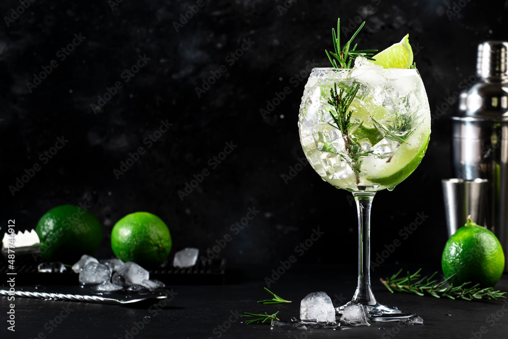Gin tonic alcoholic cocktail drink with dry gin, rosemary, tonic, lime and ice cubes in wine glass. Black bar counter background, bar tools, copy space - obrazy, fototapety, plakaty 