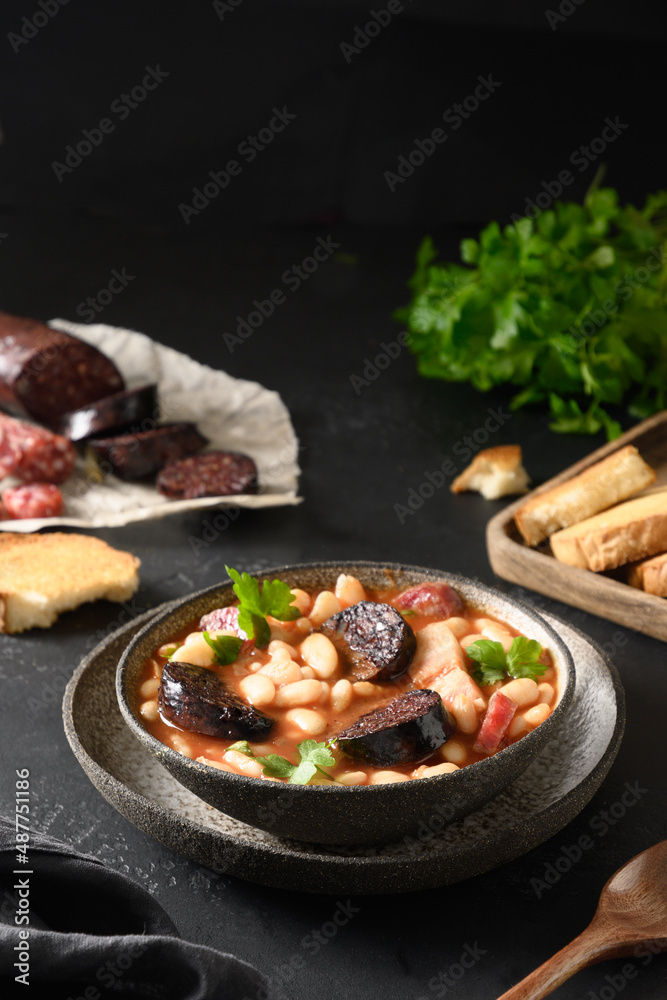 Asturiana fabada with chorizo, bacon, sausage, haricot served fresh crispy toast on black table. Vertical orientation. Tasty traditional spanish warming dinner.