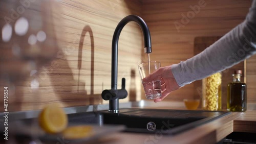 A man fills a GLASS with drinking water from a modern KITCHEN TAP. Clean and minimal interior design, side view.