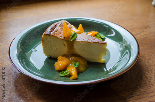 Piece of cheesecake on a green plate with peach slices and mint leaves and sea buckthorn glaze on a wooden background.