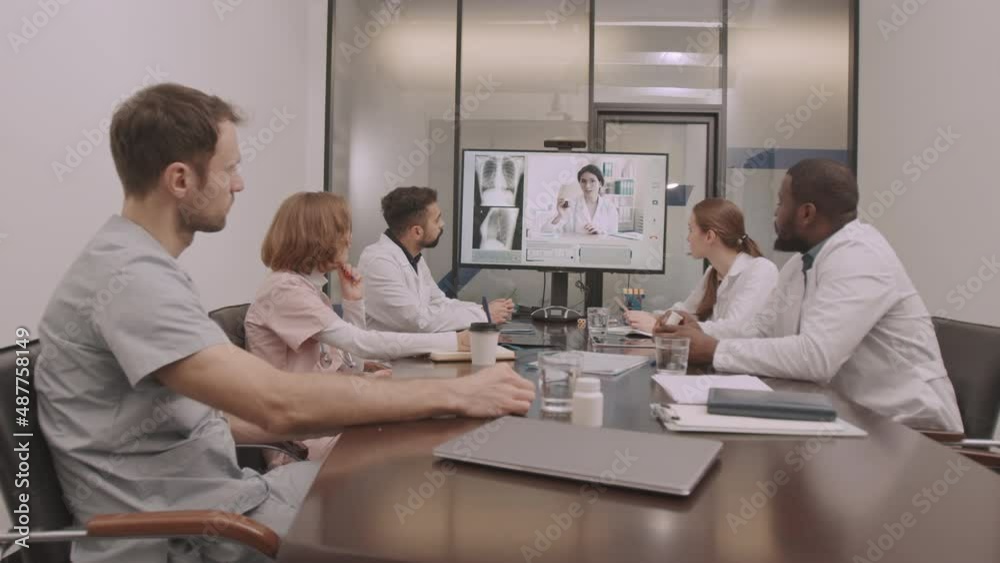 Dollying-in shot of multiethnic medical team sitting around table in conference room having online video meeting with female chief physician, discussing chest x-ray of patient Dollying-in shot of multiethnic medical team sitting around table in conference room having online video meeting with female chief physician, discussing chest x-ray of patient