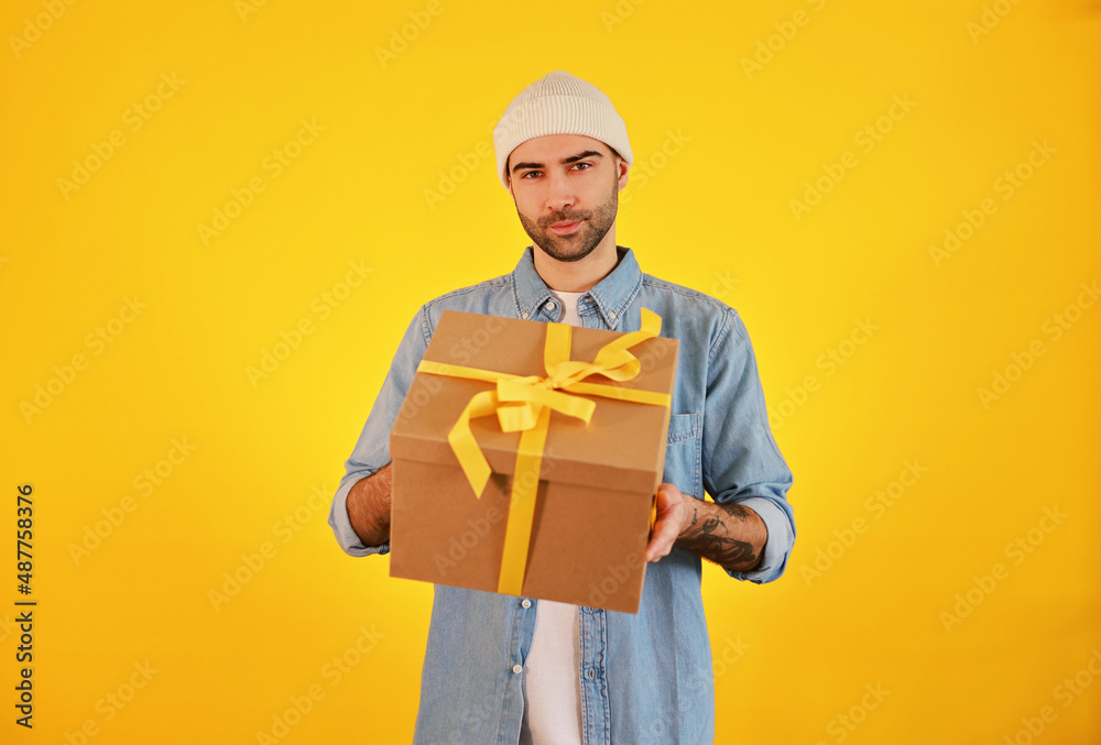 Young handsome man with big present box yellow ribbon. Women's day, 8 March, anniversary, selebration concept. Male in jeans in white hat. Young handsome man with big present box yellow ribbon. Women's day, 8 March, anniversary, selebration concept. Male in jeans in white hat.