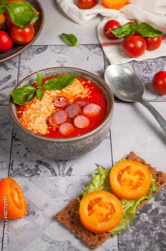 Tomato soup with smoked sausages and parmesan, on a background of gray tiles, with tomatoes and herbs