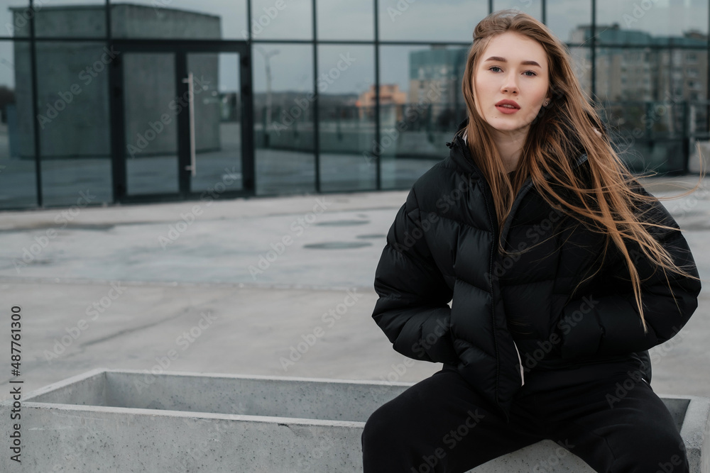 A young beautiful girl in a black short down jacket and sweatpants sitting on a concrete curb. Street style lady concept.