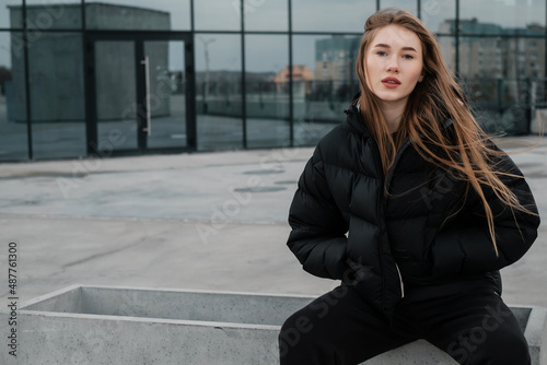 A young beautiful girl in a black short down jacket and sweatpants sitting on a concrete curb. Street style lady concept.