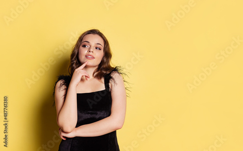Studio shot of young pretty girl with long curly hair wearing black evening dress isolated on yellow background. Concept of emotions  beauty  fashion